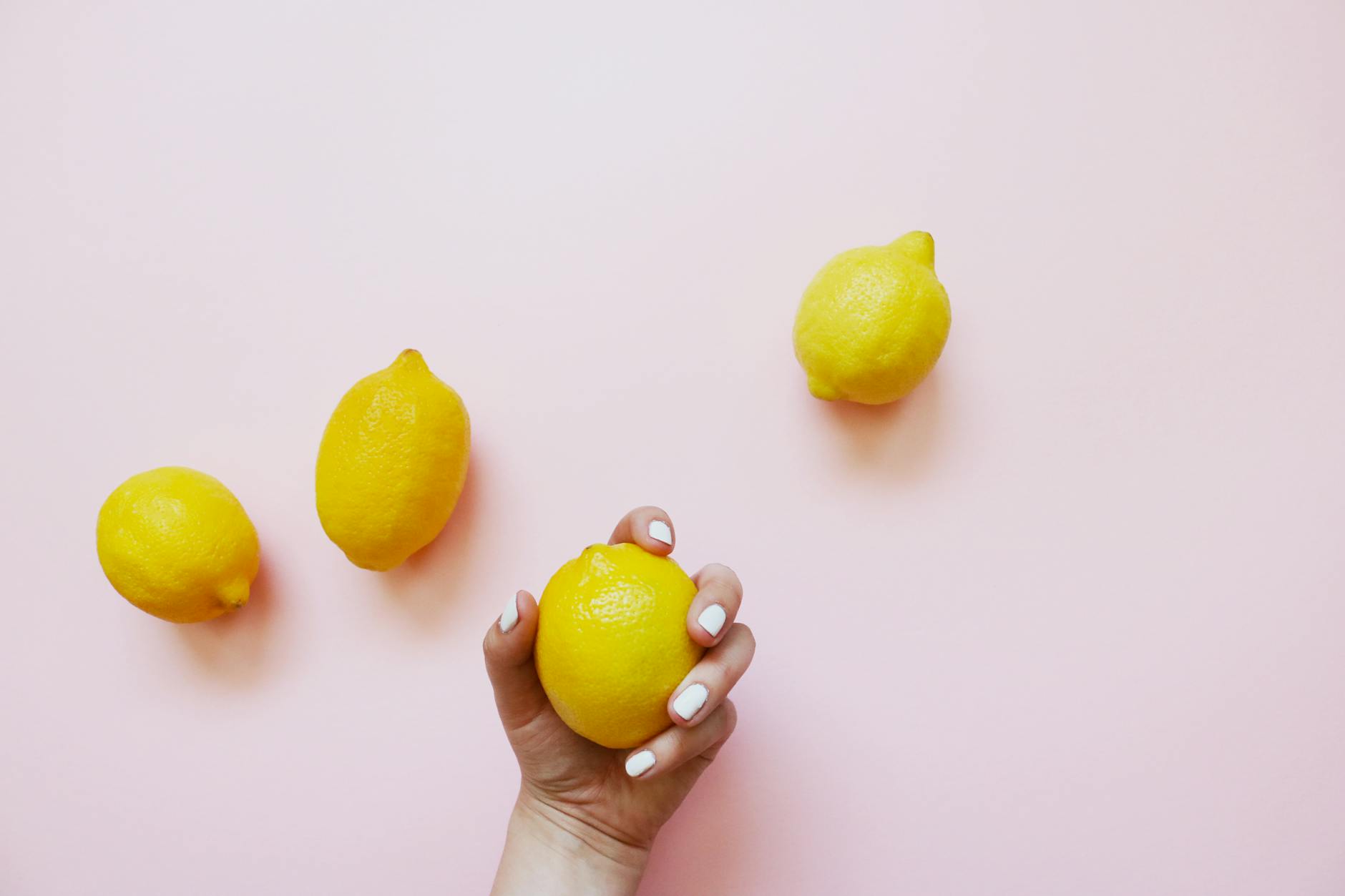 Uma mão com unhas brancas segurando um limão fresco em fundo rosa pastel