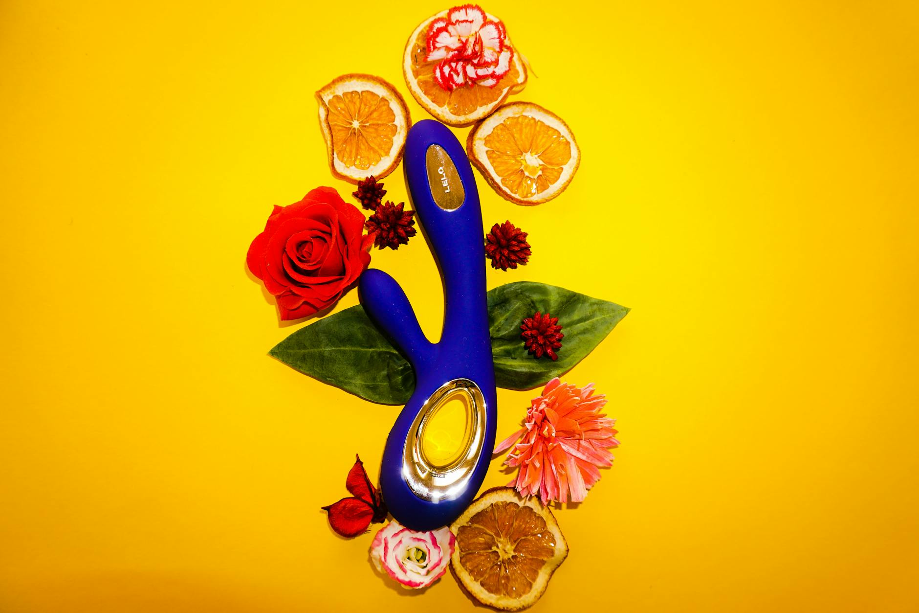 Um vibrador de sucção azul cercado por flores e fatias de laranja em um fundo amarelo