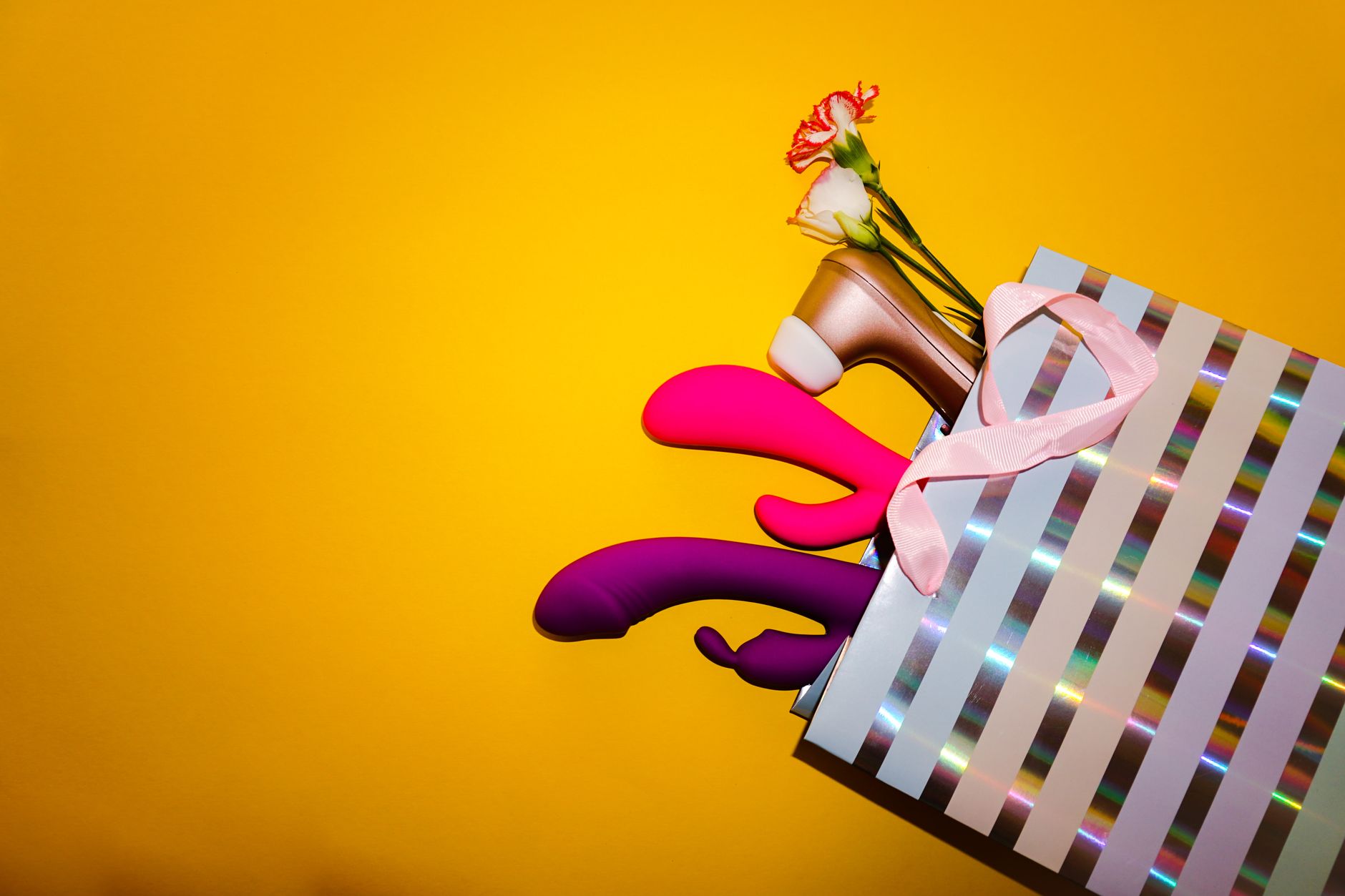 Vibradores coloridos com flores em uma bolsa de presente holográfica contra fundo amarelo vibrante.